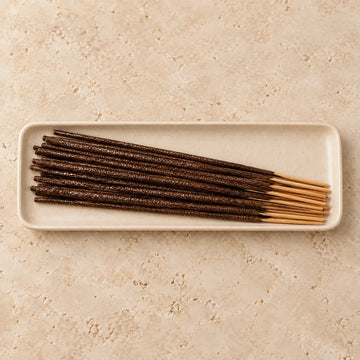 Dark brown hand-dipped incense sticks with a glossy oil sheen resting in an ivory ceramic tray.