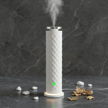 A white cylindrical humidifier with a diamond-patterned textured body, emitting a fine mist from the top. It features a glowing green indicator light and a warm white LED base, set against a dark gray marble background with decorative stones and dried leaves.