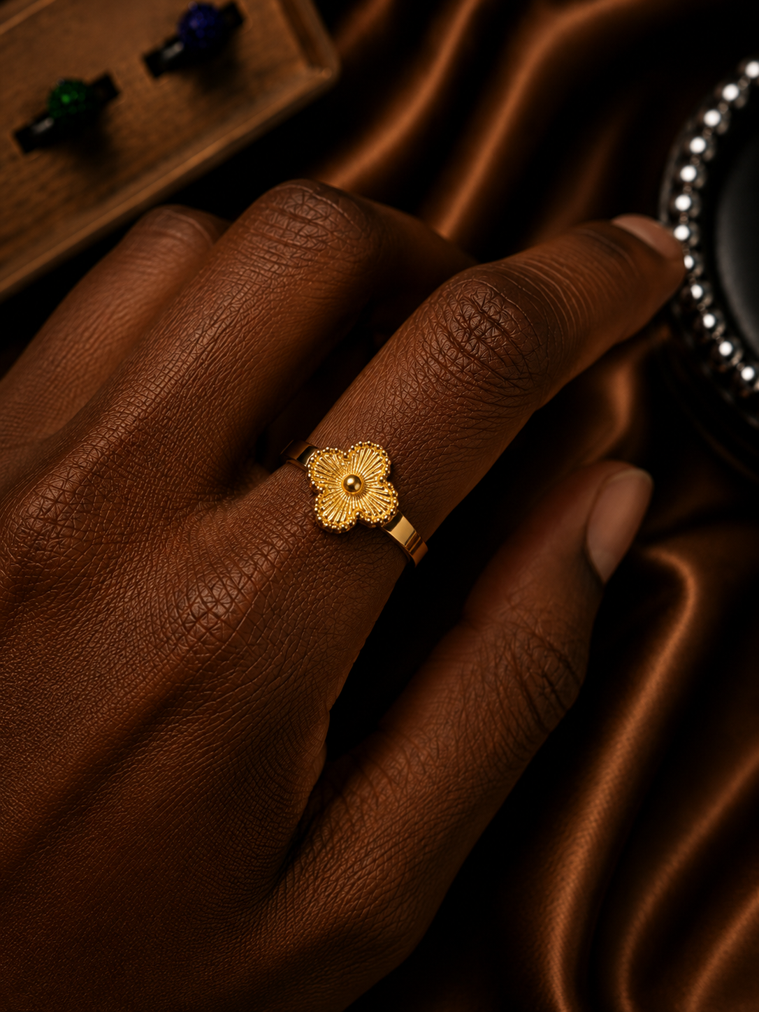 A close-up of a hand wearing a textured gold ring shaped like a stylized flower with a small central stone, set against a dark brown satin fabric background.