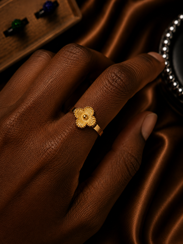 A close-up of a hand wearing a textured gold ring shaped like a stylized flower with a small central stone, set against a dark brown satin fabric background.