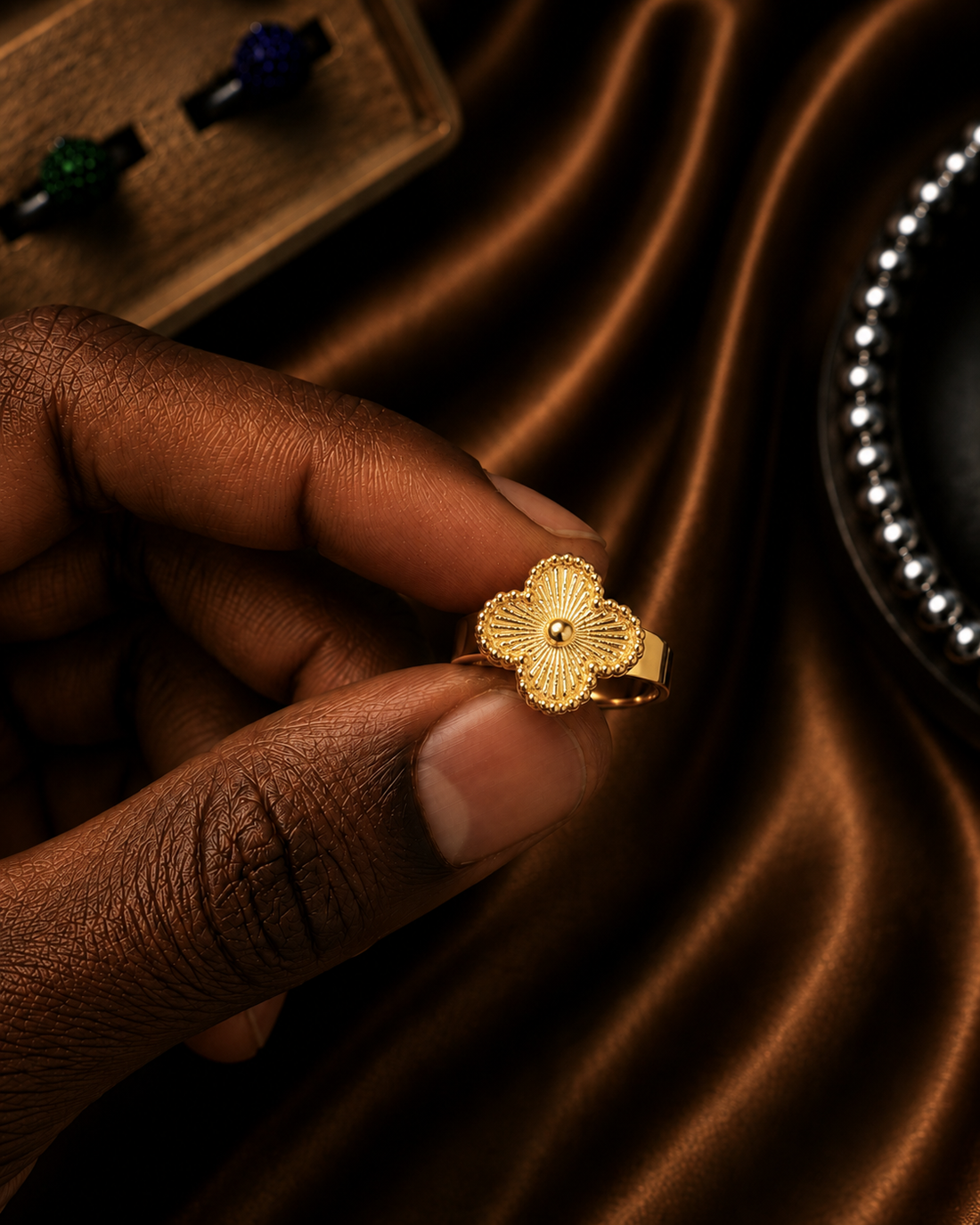 A close-up of a hand wearing a textured gold ring shaped like a stylized flower with a small central stone, set against a dark brown satin fabric background.