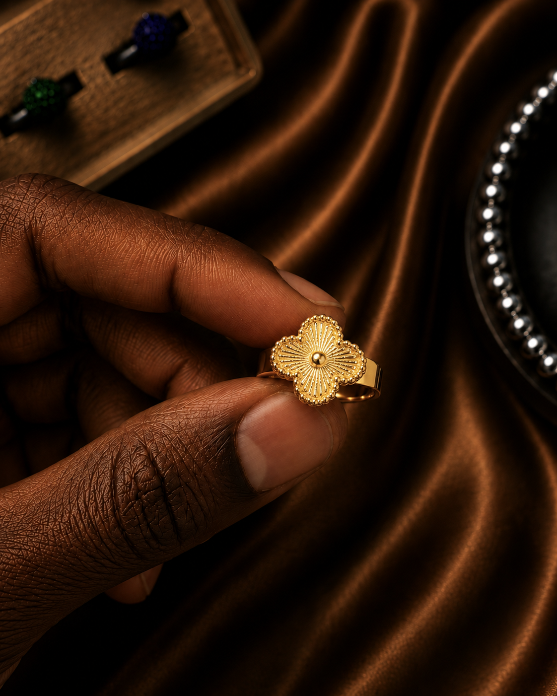 A close-up of a hand wearing a textured gold ring shaped like a stylized flower with a small central stone, set against a dark brown satin fabric background.