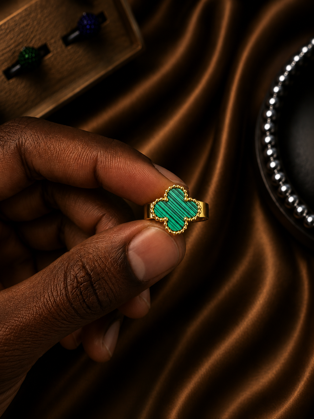 A hand holding a gold ring with a four-leaf clover-shaped green inlay, set against a luxurious brown satin fabric background.