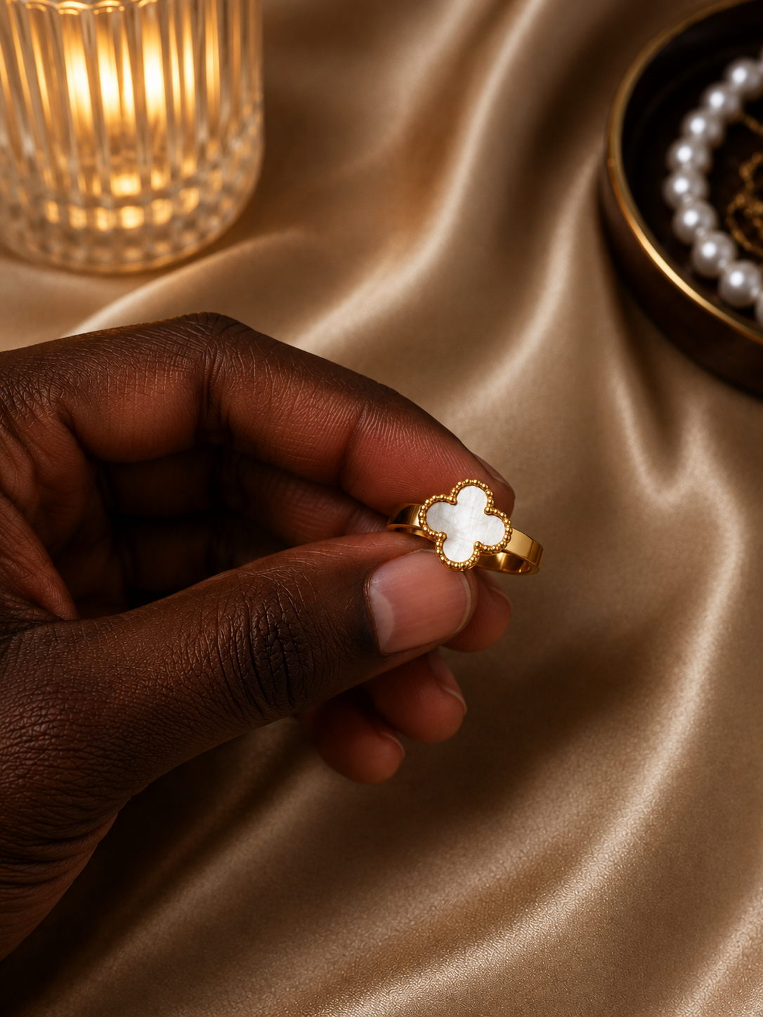 A hand holding a gold ring with a white clover-shaped inlay, set against a draped beige satin fabric with a warm lit candle and pearl jewelry in the background.