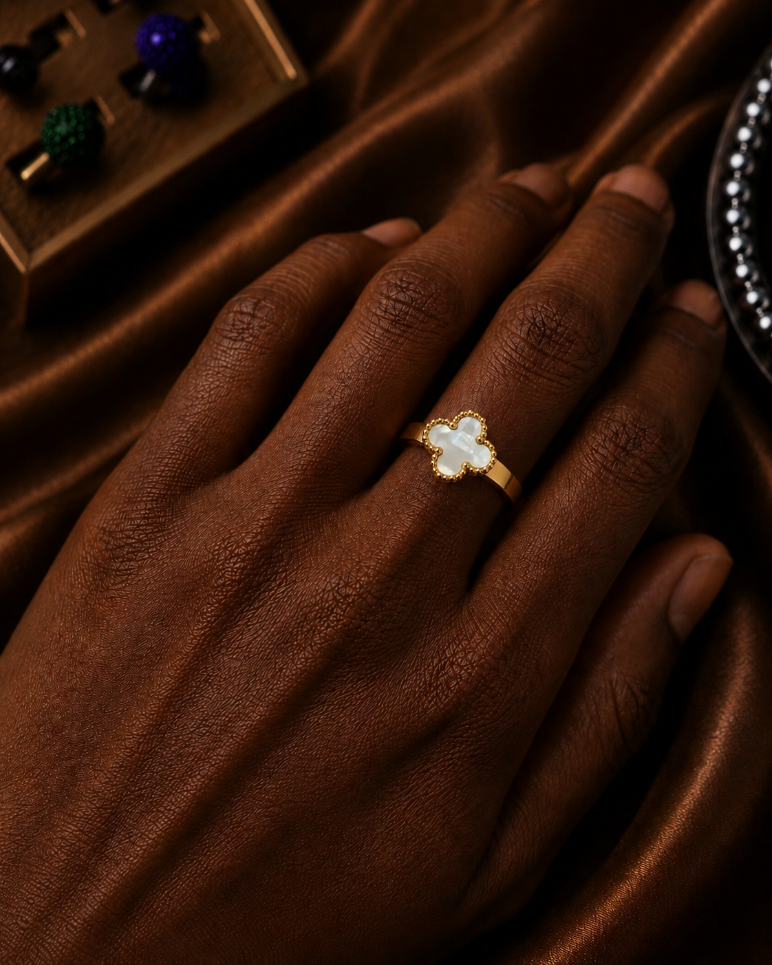 A gold ring with a white mother-of-pearl clover-shaped center stone, worn on a person's finger against a dark brown satin fabric background.